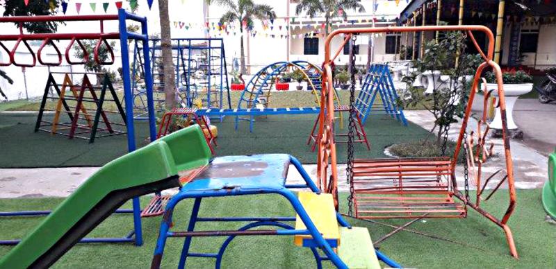 Bức tranh vẽ trường mầm non với màu sắc tươi vui và các em bé đang nô đùa trong sân trường