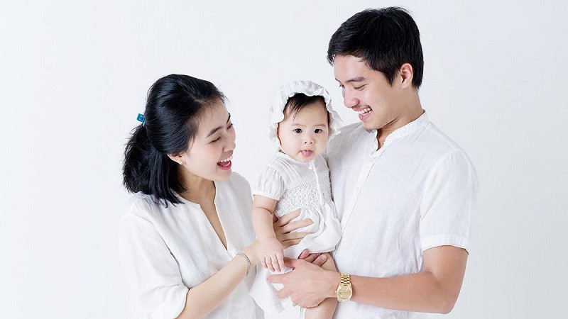 Gia đình 3 người mặc áo sơ mi trắng, đứng trên bãi cỏ, có nắng nhẹ