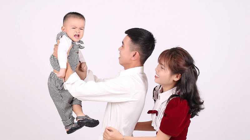 Gia đình 3 người mặc đồ trắng, ngồi trên sàn gỗ chụp ảnh studio