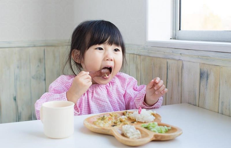 Bé trai ngồi bàn ăn tập xúc thìa, minh họa kỹ năng tự ăn uống.